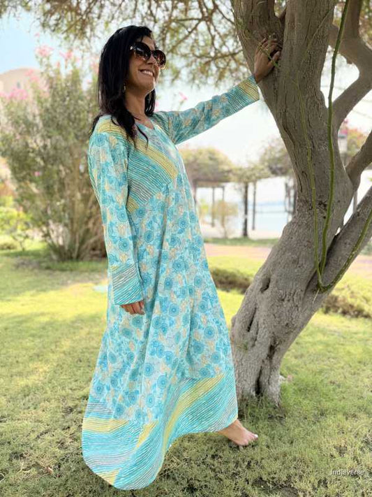 Woman in a long, light blue dress with yellow accents standing next to a tree outdoors.
