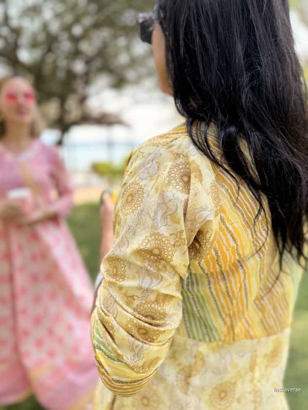 Person wearing a patterned yellow and green outfit with another person in a pink dress in the background.