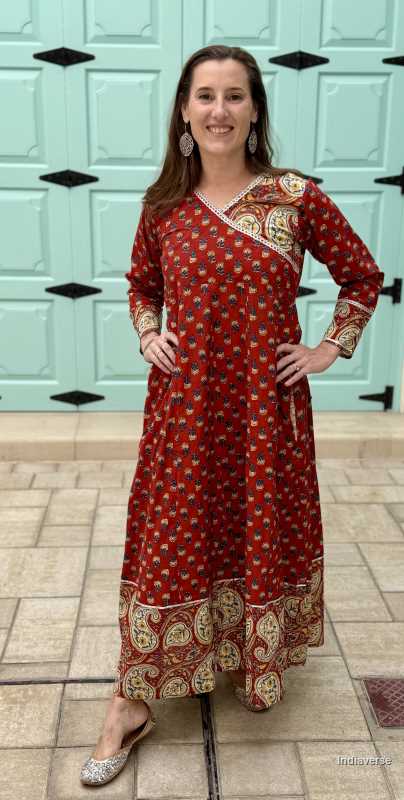 Woman wearing long maroon red colored wrap dress with paisley print border and silver border details around the next and sleeves in front a of a light blue wooden door