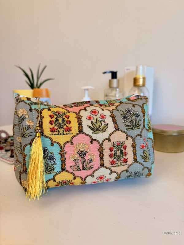 A rectangular cosmetic pouch with a colorful patchwork floral pattern and a gold tassel zipper pull, displayed on a tabletop with skincare items in the background | Arabesque