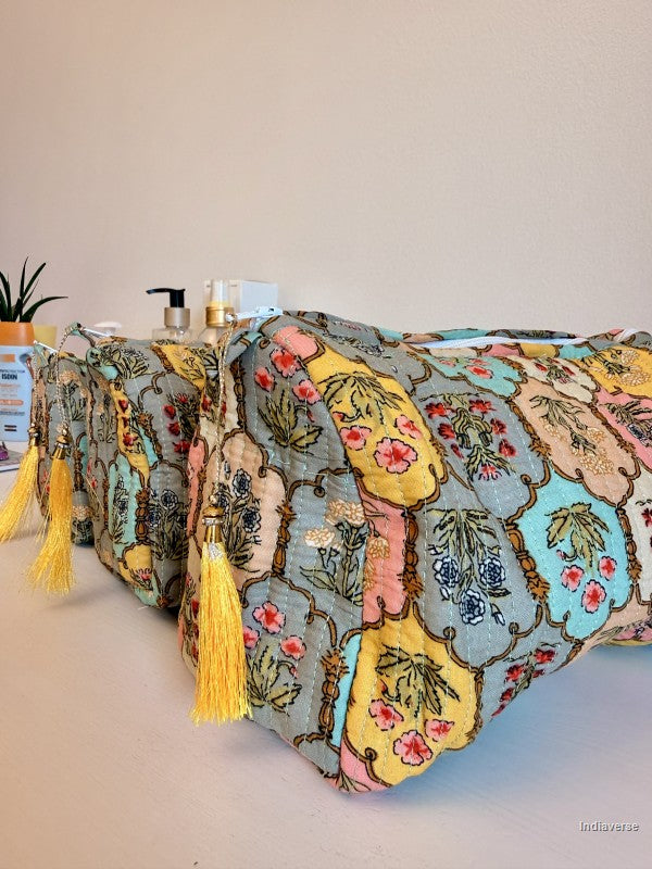 A close-up of a patterned textile product draped over a surface, featuring a multicolor floral and ornamental print in yellow, pink, blue, beige, and green, with prominent yellow tassels hanging down. The background is a plain beige wall with a few small decorative items on a shelf | Arabesque