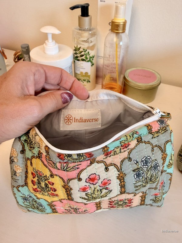 A hand holds open a patterned makeup bag with a visible Indiaverse brand label inside. The bag has a multicolor floral and ornamental print on a pale blue-gray background, with a white zipper trim | Arabesque