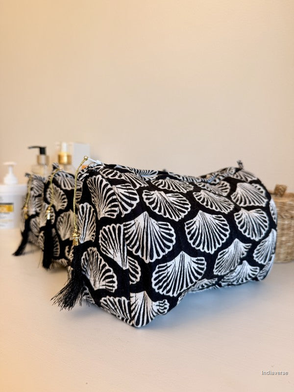 A black and white patterned cosmetic bag with tassel zipper pulls, displayed on a light beige surface with toiletry bottles in the background | Black Sea Shell