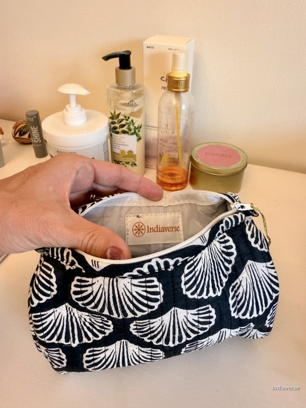 A hand holding open a navy and white ginkgo leaf patterned cosmetic pouch, showing the Indiaversa brand label inside. The pouch is set on a light surface with various toiletries in the background | Black Sea Shell
