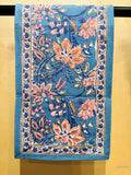 A rectangular blue table runner with an ornate floral pattern and decorative border, hanging against a warm yellow wall.