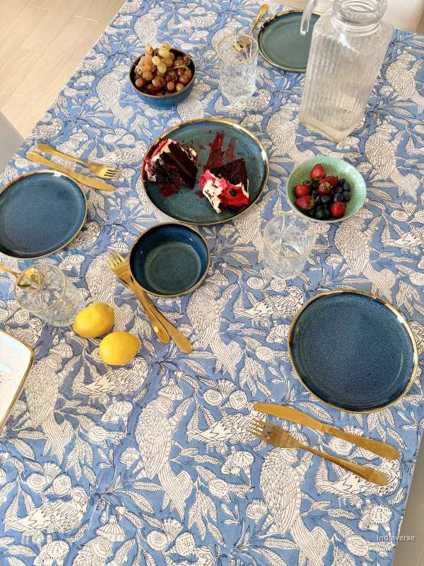 Dining table setting with blue plates, gold cutlery, and fruit on a decorative tablecloth.