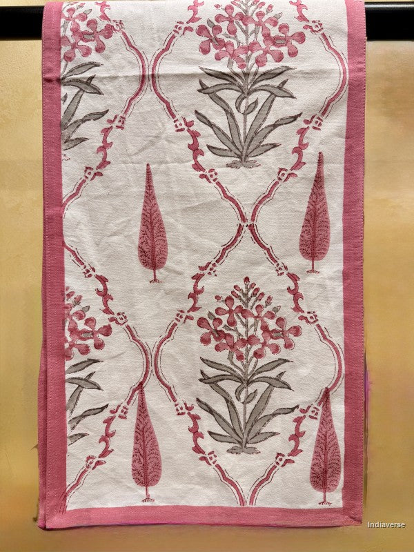 A rectangular textile panel or scarf with a cream background, featuring a repeating pink and gray floral trellis pattern and a solid pink border, shown hanging against a tan wall.