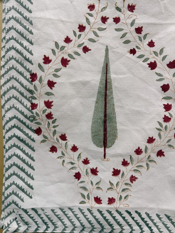 A close-up of a white textile featuring a central tree motif framed by a circular wreath of red flowers and green leaves, with decorative green and white patterned borders along the left and bottom edges.