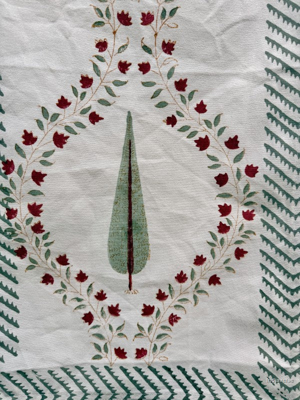 A close-up of a white textile featuring a central green leaf motif surrounded by a wreath of red flowers and green leaves, with green geometric border patterns along the edges.