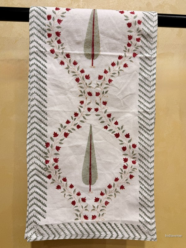 A white cotton kitchen towel with a red floral vine border and gray-green teardrop motifs, hanging on a black rod against a beige wall.