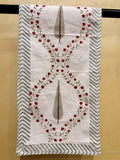 A white cotton kitchen towel with a red floral vine border and gray-green teardrop motifs, hanging on a black rod against a beige wall.