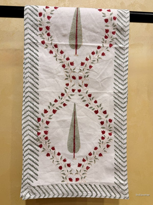 A white cotton kitchen towel with a red floral vine border and gray-green teardrop motifs, hanging on a black rod against a beige wall.