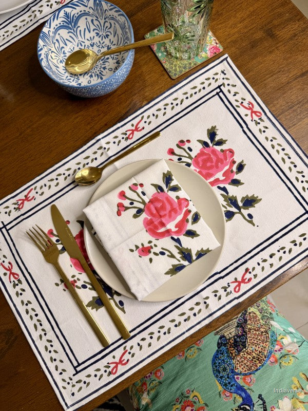 A placemat with rose design arranged on a brown wooden table with a beige color plate and a rose design napkin on top. 