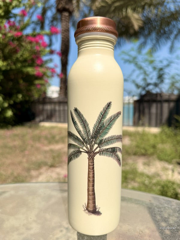 beige copper water bottle with single palm tree design standing on a glass table inside a garden with a palm tree in the background