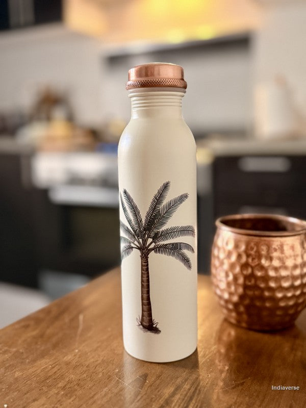 beige copper water bottle with single palm tree design standing on a brown table in a kitchen with a copper cup in the background