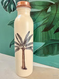 beige copper water bottle with single palm tree design standing in front of a green leaf wallpaper