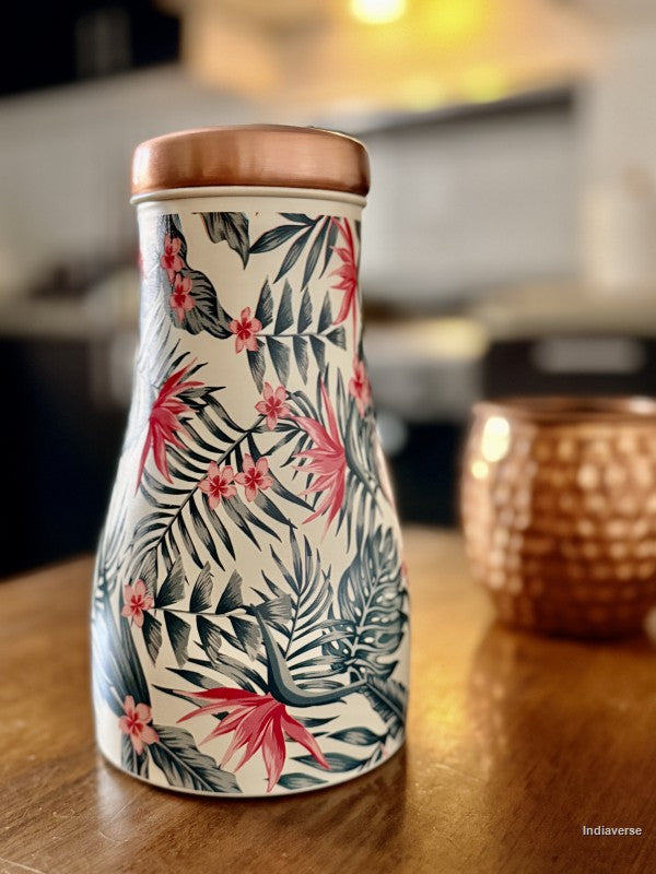 Copper water jar with tropical leaf and birds of paradise design sitting on a brown table in a kitchen with a copper cup in the background