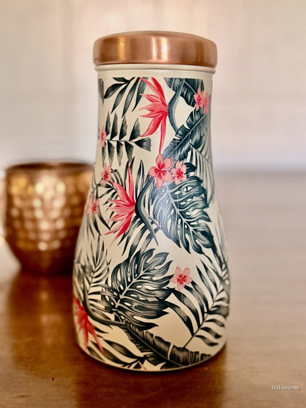 Copper water jar with tropical leaf and birds of paradise design sitting on a brown table with a copper cup in the background