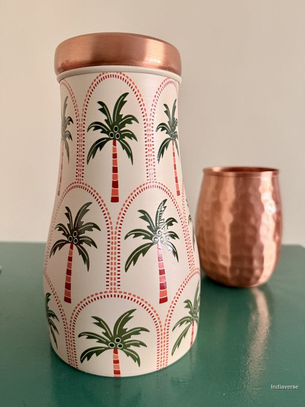 A decorative copper water jar with geometric palm tree design is sitting on a green surface with a copper mug in the background