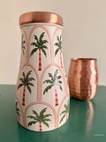 A decorative copper water jar with geometric palm tree design is sitting on a green surface with a copper mug in the background