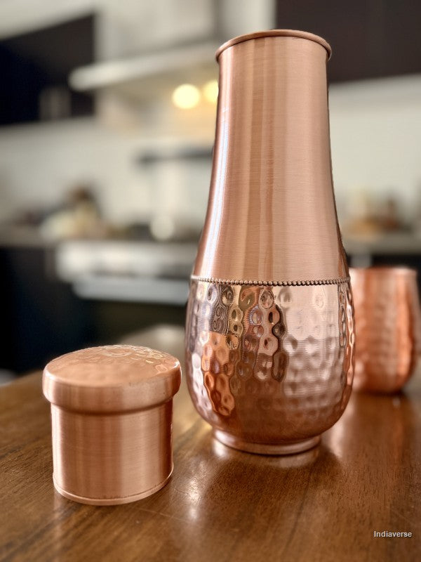 tall pure copper water jar with hammered bottom and smooth copper body sitting on a brown table with its lid next to it