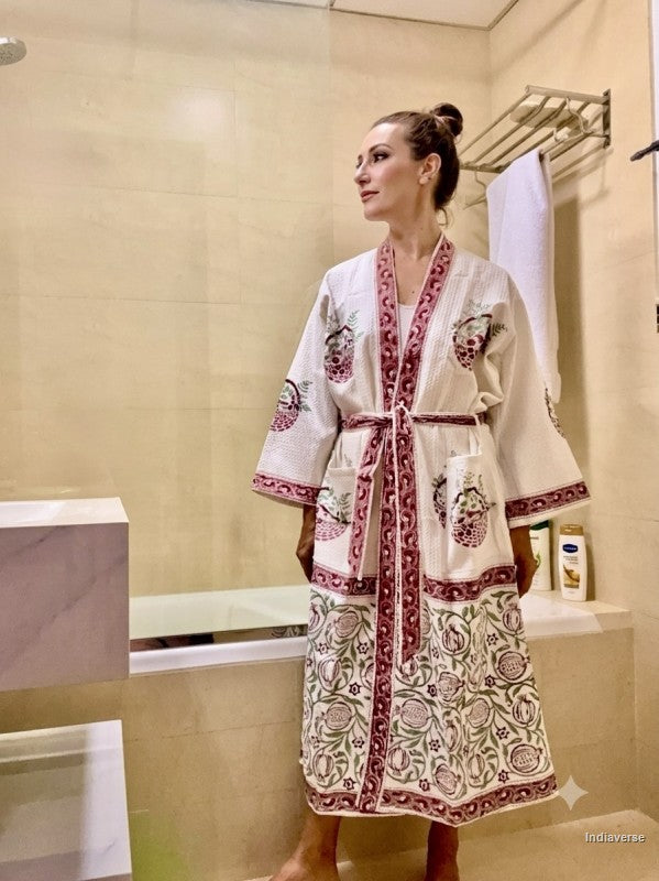 A woman wearing a cream-colored cotton robe with red and green floral embroidery and a red patterned border, standing in a tiled bathroom.