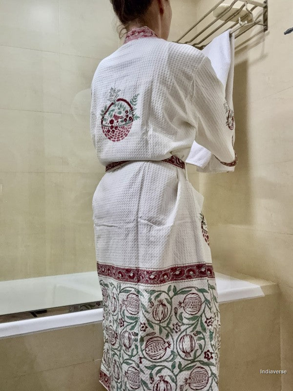 A woman viewed from behind wearing a white waffle-knit robe with red and green floral embroidery, standing in a tiled bathroom.