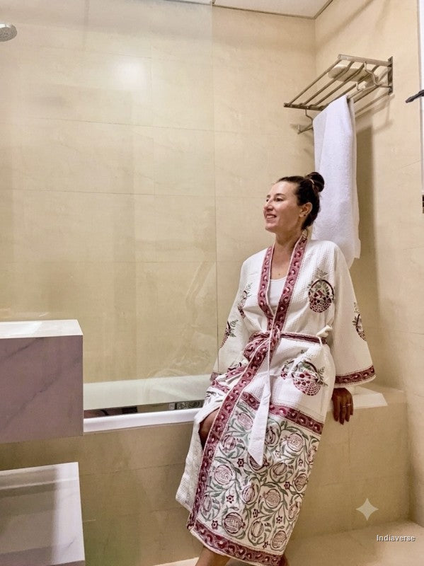 A woman wearing a white cotton robe with red and green floral embroidery and a red border, standing in a beige tiled bathroom.