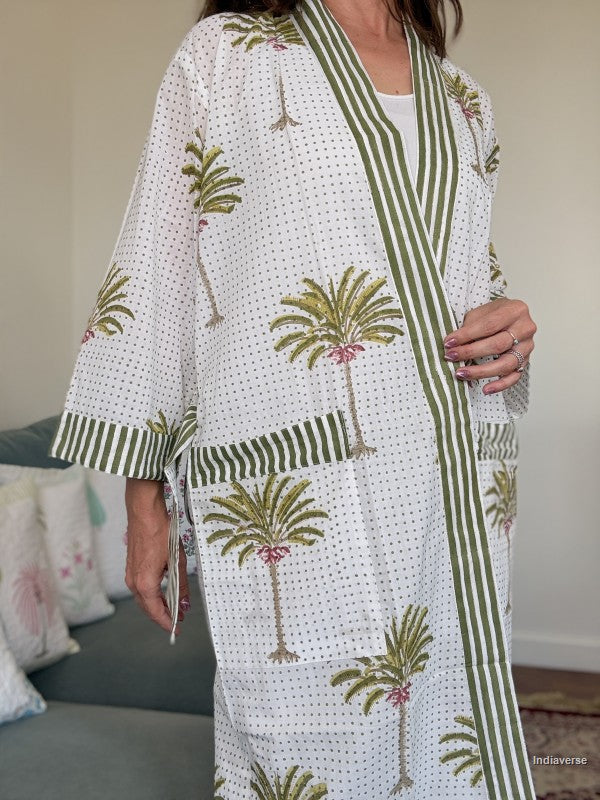 A woman wearing a white kimono-style robe with green striped trim and green palm tree prints, standing indoors in a living room setting.