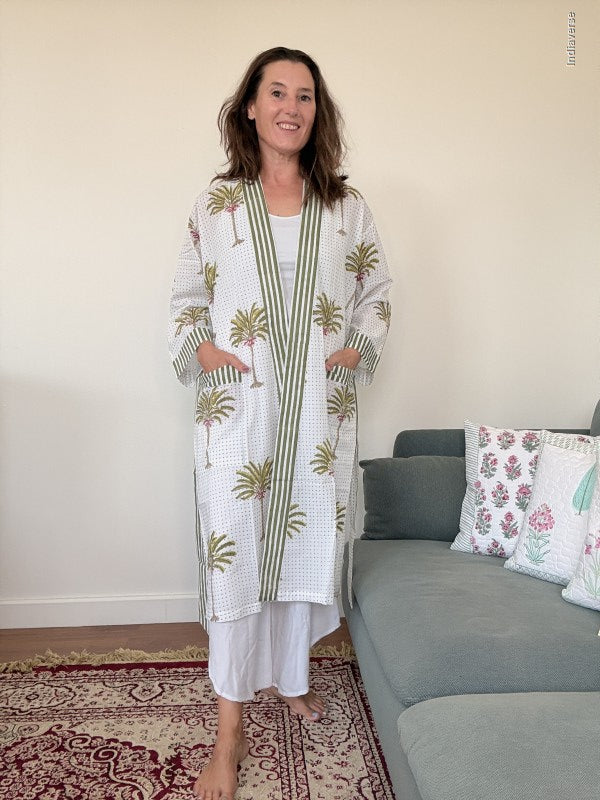 A woman stands barefoot in a living room wearing a white long robe with green palm tree prints and green striped borders. The robe has pockets and is worn over white pants.