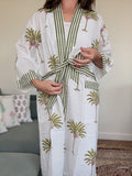 A woman wearing a white cotton robe with green and white striped trim, green palm tree prints, and small pink floral accents, standing in a living room setting.