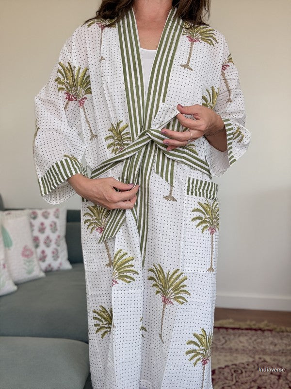 A woman wearing a white cotton robe with green and white striped trim, green palm tree prints, and small pink floral accents, standing in a living room setting.
