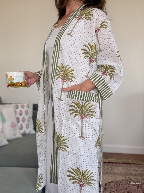 A woman wearing a white open-front robe with green striped borders and green palm tree prints, holding a small colorful mug in a living room setting.