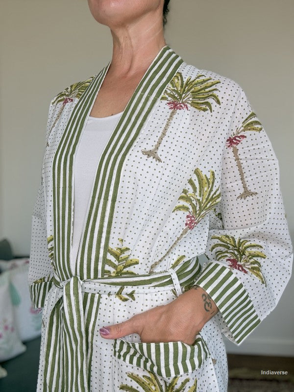 A woman wearing a white cotton robe with green vertical stripes, green palm tree prints, and small pink floral accents. The robe features a tie belt and a front pocket.