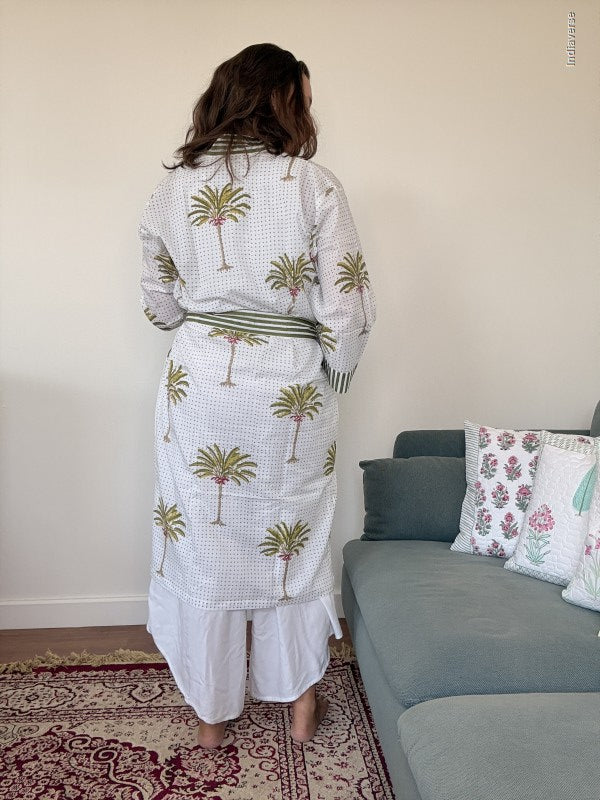 A woman stands with her back to the camera wearing a white checkered long-sleeve robe with a green and gold palm tree pattern, paired with white pants. She is standing in a living room next to a gray sofa with patterned pillows.