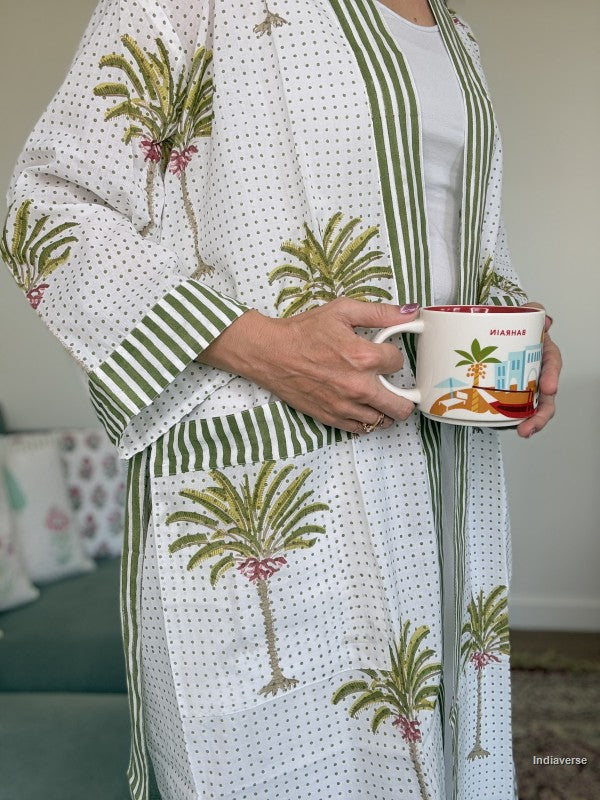 A person wearing a white kimono-style robe with green striped trim and green palm tree prints, holding a white ceramic mug with a colorful beach illustration.