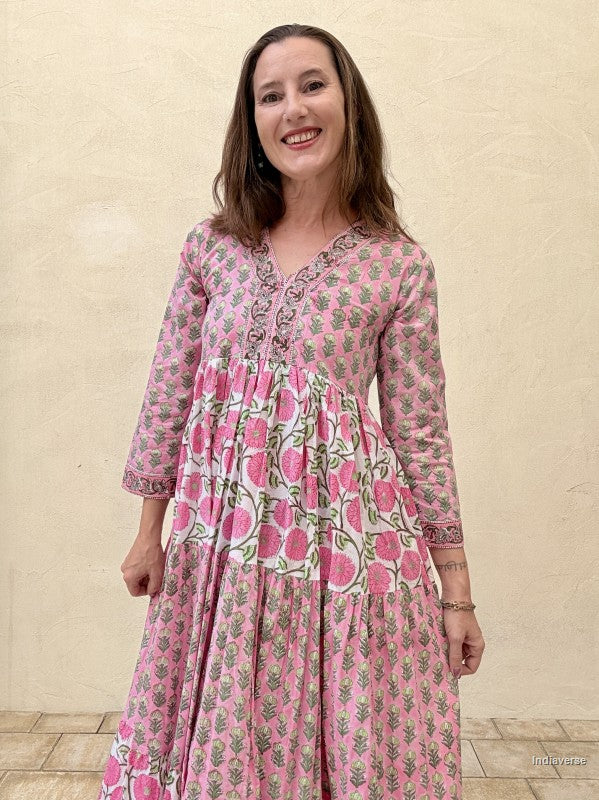 Woman wearing a long cotton maxi dress with traditional small flower design and big pink poppy motif standing against a beige wall