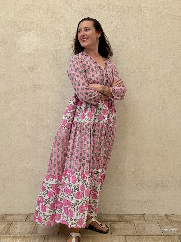 Woman wearing a long cotton maxi dress with traditional small flower design and big pink poppy motif standing against a beige wall