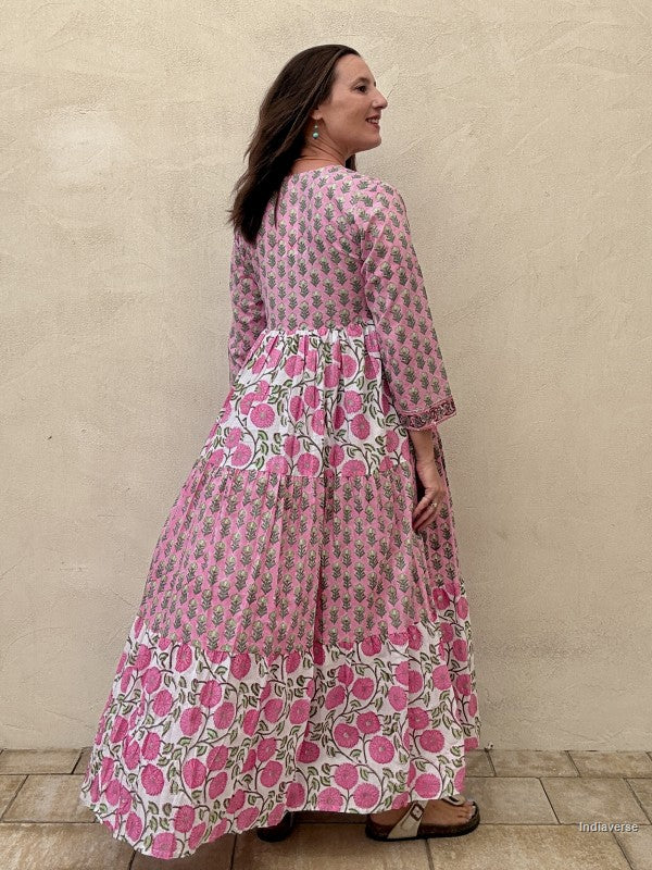 Backside view of a woman wearing a long cotton maxi dress with traditional small flower design and big pink poppy motif standing against a beige wall