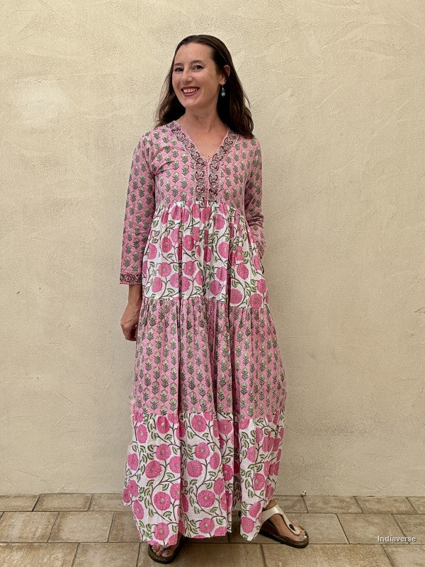 Woman wearing a long cotton maxi dress with traditional small flower design and big pink poppy motif standing against a beige wall