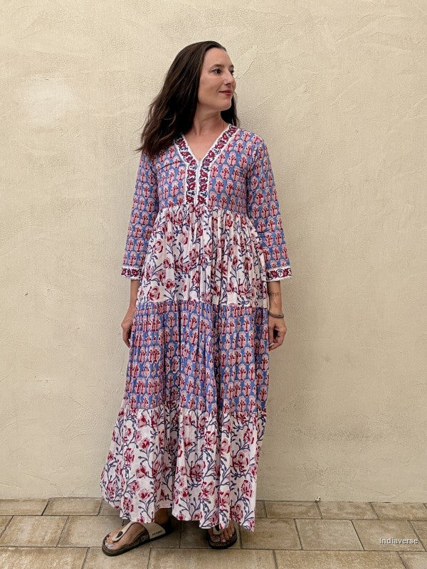 A woman modeling a long tiered maxi dress with a blue, pink, and white floral print, standing against a textured beige stucco wall.