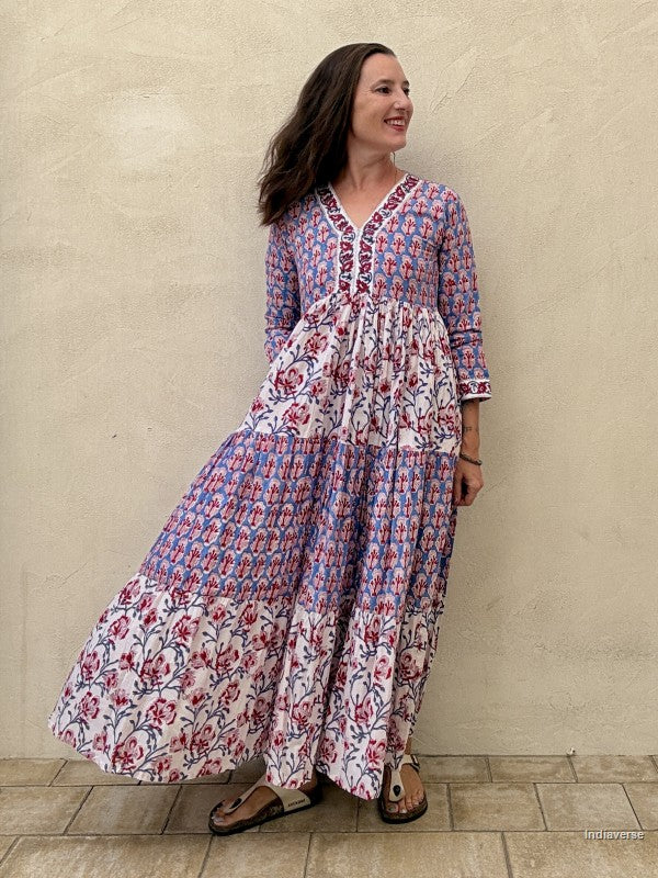 A woman wearing a long tiered maxi dress with a blue, red, and white floral block print pattern, standing against a textured beige stucco wall.