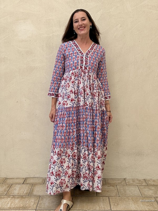 A woman modeling a long-sleeve maxi dress with a blue, red, and white floral and geometric pattern, standing against a textured beige stucco wall.