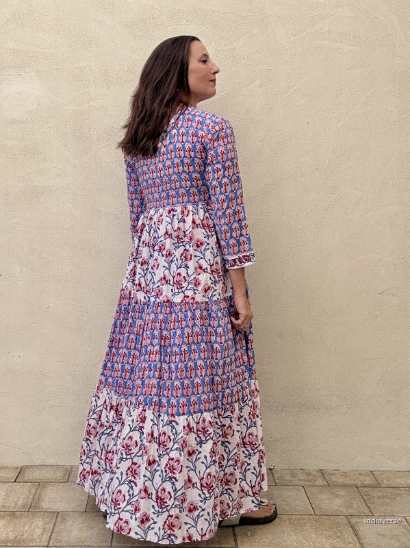 A woman wearing a long tiered maxi dress with a blue and pink floral block print, shown from a side-back angle against a textured beige stucco wall.