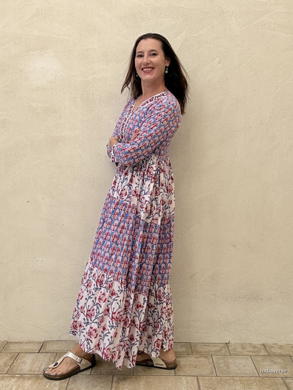 A woman modeling a long-sleeve maxi dress with a blue, pink, and white floral pattern, standing against a textured beige stucco wall.