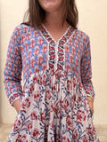 A woman wearing a tiered cotton dress with a red, blue, and white block print floral pattern, featuring a V-neckline and long sleeves.