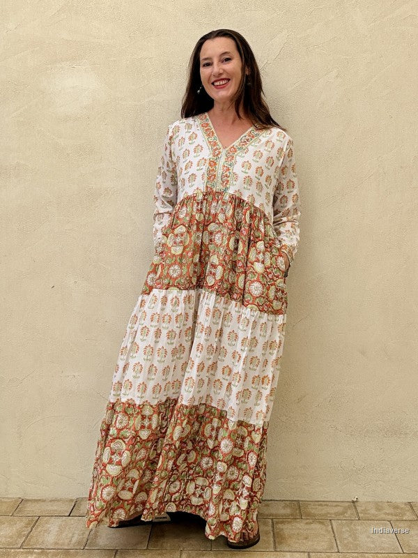 A Woman wearing a long maxi dress made from cotton with floral and palm tree print motif standing against a beige wall