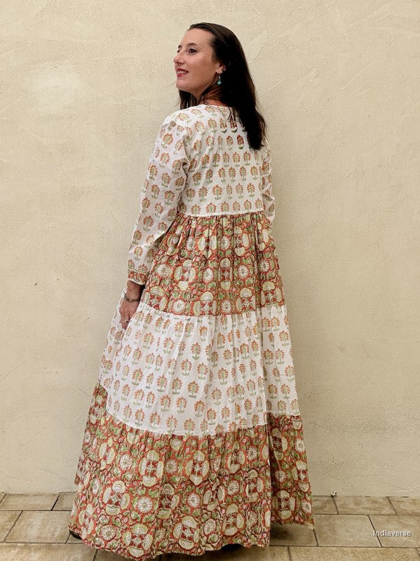 View of the backside of a woman wearing a long maxi dress made from cotton with floral and palm tree print motif standing against a beige wall