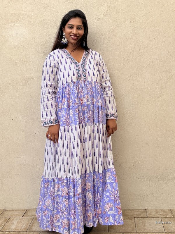 Woman standing in front of a beige wall wearing a beautiful long cotton dress with floral and cypress prints in aquamarine blue tones