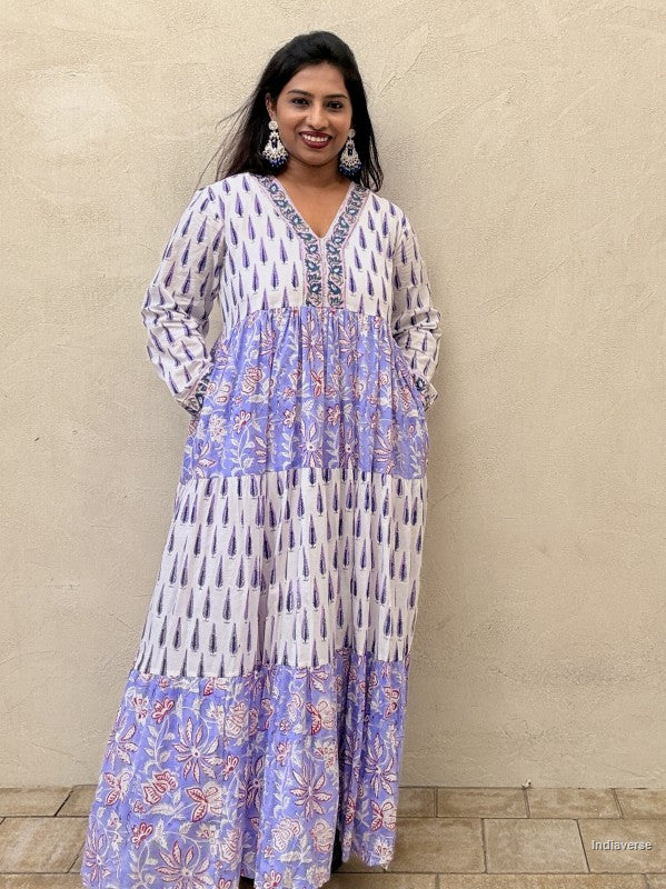 Woman standing in front of a beige wall wearing a beautiful long cotton dress with floral and cypress prints in aquamarine blue tones